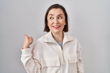 Foto de Mujer hispana de mediana edad de pie sobre un fondo aislado sonriendo con la cara feliz mirando y señalando hacia un lado con el pulgar hacia arriba. - Imagen libre de derechos
