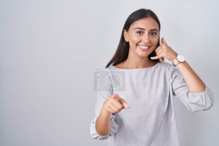 Foto de Young hispanic woman standing over white background smiling doing talking on the telephone gesture and pointing to you. call me. - Imagen libre de derechos