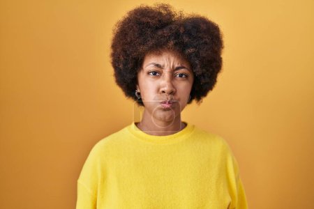 Téléchargez les photos : Young african american woman standing over yellow background puffing cheeks with funny face. mouth inflated with air, crazy expression. - en image libre de droit