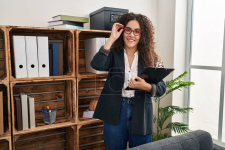 Photo for Young beautiful hispanic woman business worker smiling confident holding clipboard at office - Royalty Free Image