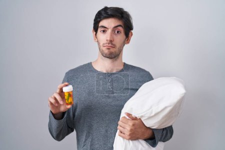Foto de Joven hombre hispano usando pijama abrazando almohada sosteniendo pastillas sin idea y expresión confusa. concepto de duda. - Imagen libre de derechos