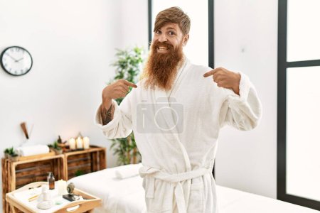 Foto de Pelirrojo con barba larga usando albornoz en el spa wellness mirando confiado con sonrisa en la cara, señalándose con los dedos orgullosos y felices. - Imagen libre de derechos