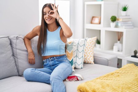 Téléchargez les photos : Jeune femme brune assise sur le canapé à la maison faisant un geste correct avec la main souriante, les yeux regardant à travers les doigts avec un visage heureux. - en image libre de droit