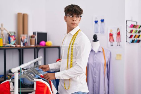 Téléchargez les photos : Jeune adolescent hispanique tailleur tenant des vêtements sur rack au magasin de tailleur - en image libre de droit
