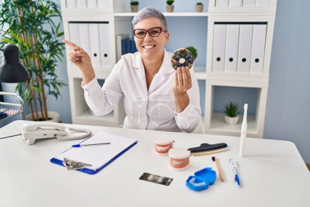 Photo for Middle age dentist woman holding chocolate doughnut smiling with an idea or question pointing finger up with happy face, number one - Royalty Free Image