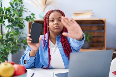 Foto de African american woman with braided hair holding smartphone showing blank screen with open hand doing stop sign with serious and confident expression, defense gesture - Imagen libre de derechos