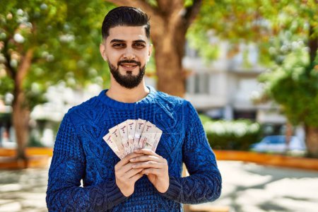 Téléchargez les photos : Jeune homme arabe souriant confiant tenant des billets riyal au parc - en image libre de droit