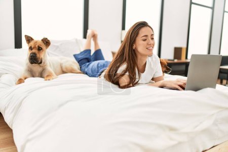 Téléchargez les photos : Jeune femme hispanique utilisant un ordinateur portable couché sur le lit avec des chiens dans la chambre - en image libre de droit