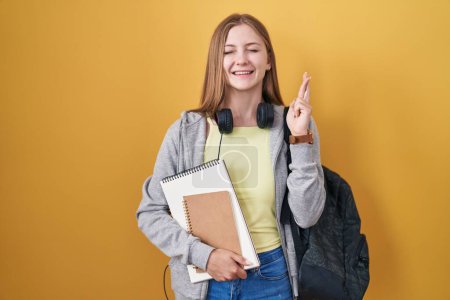 Foto de Young caucasian woman wearing student backpack and holding books gesturing finger crossed smiling with hope and eyes closed. luck and superstitious concept. - Imagen libre de derechos
