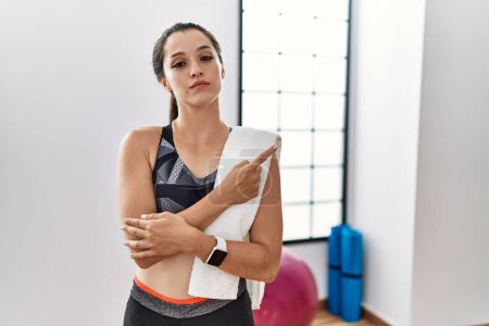 Téléchargez les photos : Jeune femme brune portant des vêtements de sport et une serviette à la salle de gym pointant du doigt vers le côté montrant la publicité, visage sérieux et calme - en image libre de droit