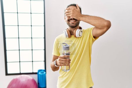 Photo for Young hispanic man wearing sportswear and drinking water at the gym smiling and laughing with hand on face covering eyes for surprise. blind concept. - Royalty Free Image