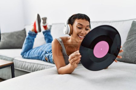 Foto de Joven mujer hispana escuchando música sosteniendo un disco de vinilo tumbado en un sofá en casa - Imagen libre de derechos