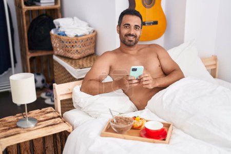 Téléchargez les photos : Young hispanic man using smartphone having breakfast sitting on bed at bedroom - en image libre de droit