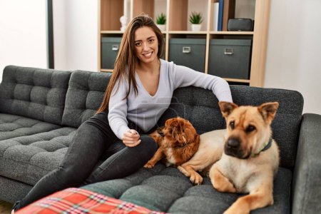 Foto de Joven mujer hispana sonriendo confiada sentada en sofá con perros en casa - Imagen libre de derechos