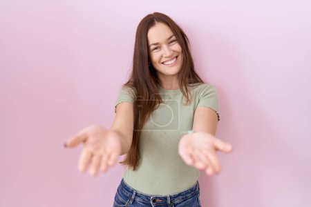 Foto de Hermosa mujer morena de pie sobre fondo rosa sonriente alegre ofreciendo manos dando asistencia y aceptación. - Imagen libre de derechos