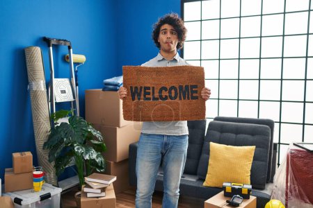 Photo for Hispanic man with curly hair holding welcome doormat making fish face with mouth and squinting eyes, crazy and comical. - Royalty Free Image