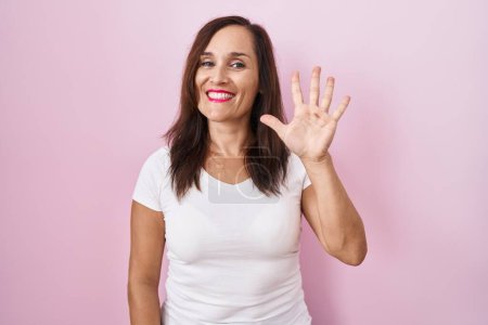 Foto de Middle age brunette woman standing over pink background showing and pointing up with fingers number five while smiling confident and happy. - Imagen libre de derechos