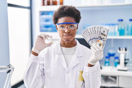 Foto de Mujer afroamericana trabajando en laboratorio científico sosteniendo dólares soplando mejillas con cara graciosa. boca inflada con aire, captación de aire. - Imagen libre de derechos