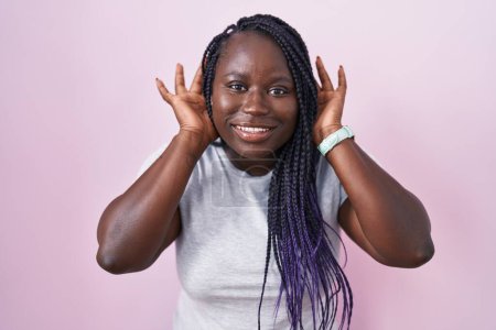 Foto de Young african woman standing over pink background trying to hear both hands on ear gesture, curious for gossip. hearing problem, deaf - Imagen libre de derechos