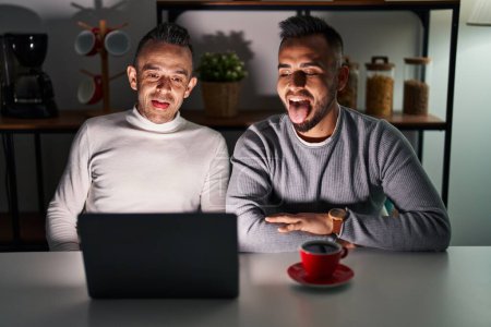 Foto de Pareja homosexual usando computadora portátil sacando la lengua feliz con expresión divertida. concepto de emoción. - Imagen libre de derechos