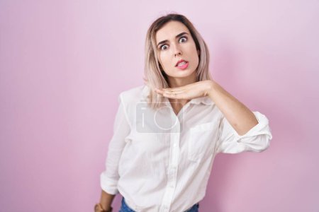 Foto de Young beautiful woman standing over pink background cutting throat with hand as knife, threaten aggression with furious violence - Imagen libre de derechos
