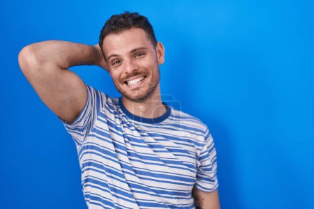 Foto de Joven hombre hispano de pie sobre fondo azul sonriendo confiado tocando el cabello con la mano hacia arriba gesto, posando atractivo y de moda - Imagen libre de derechos