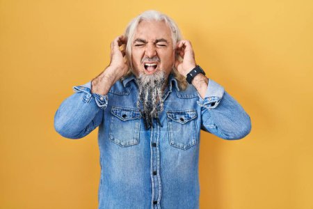 Téléchargez les photos : Middle age man with grey hair standing over yellow background covering ears with fingers with annoyed expression for the noise of loud music. deaf concept. - en image libre de droit