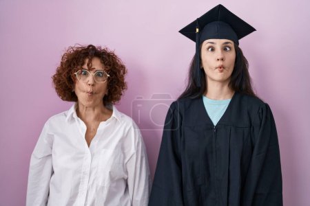 Téléchargez les photos : Hispanic mother and daughter wearing graduation cap and ceremony robe making fish face with lips, crazy and comical gesture. funny expression. - en image libre de droit