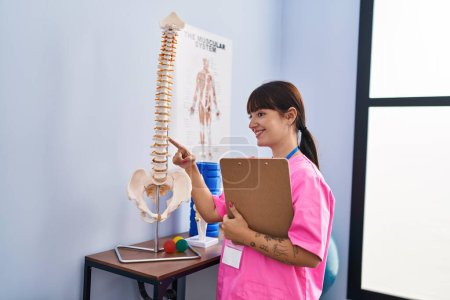 Téléchargez les photos : Jeune belle femme hispanique physiothérapeute pointant vers le modèle anatomique de colonne vertébrale tenant presse-papiers à la clinique de réadaptation - en image libre de droit