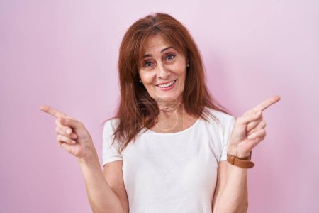 Téléchargez les photos : Femme d'âge moyen debout sur fond rose souriant confiant pointant avec les doigts vers différentes directions. espace de copie pour la publicité - en image libre de droit