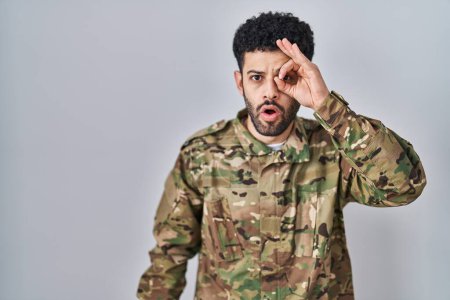 Foto de Hombre árabe vistiendo uniforme camuflaje del ejército haciendo un gesto bien sorprendido con la cara sorprendida, los ojos mirando a través de los dedos. expresión incrédula. - Imagen libre de derechos