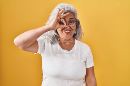 Photo for Middle age woman with grey hair standing over yellow background doing ok gesture with hand smiling, eye looking through fingers with happy face. - Royalty Free Image