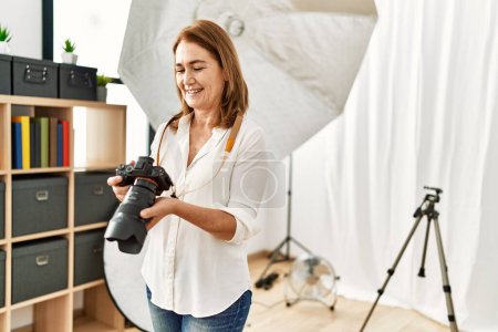 Foto de Edad media mujer caucásica fotógrafo sonriendo confiado sosteniendo cámara profesional en estudio de fotografía - Imagen libre de derechos