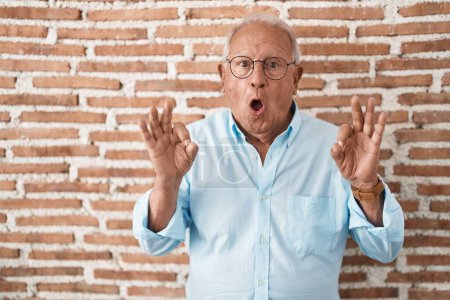 Foto de Hombre mayor con el pelo gris de pie sobre la pared de ladrillos mirando sorprendido y sorprendido haciendo ok símbolo de aprobación con los dedos. expresión loca - Imagen libre de derechos