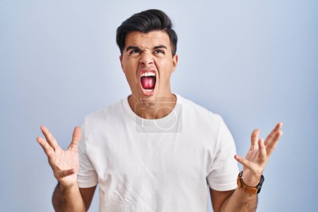 Photo for Hispanic man standing over blue background crazy and mad shouting and yelling with aggressive expression and arms raised. frustration concept. - Royalty Free Image