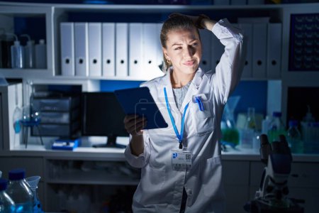 Foto de Beautiful blonde woman working at scientist laboratory late at night confuse and wondering about question. uncertain with doubt, thinking with hand on head. pensive concept. - Imagen libre de derechos