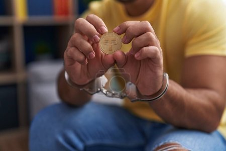 Photo for Young hispanic man criminal holding tether crypto currency wearing handcuffs at home - Royalty Free Image