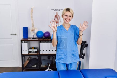 Photo for Young physiotherapist woman working at pain recovery clinic showing and pointing up with fingers number nine while smiling confident and happy. - Royalty Free Image