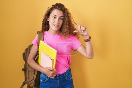 Photo for Young caucasian woman wearing student backpack and holding books smiling and confident gesturing with hand doing small size sign with fingers looking and the camera. measure concept. - Royalty Free Image