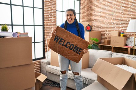 Photo for Young hispanic woman holding welcome doormat at new home smiling and laughing hard out loud because funny crazy joke. - Royalty Free Image