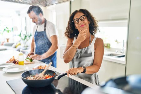 Téléchargez les photos : Couple d'âge moyen cuisiner des aliments méditerranéens à la maison visage sérieux penser à la question avec la main sur le menton, réfléchi sur l'idée déroutante - en image libre de droit