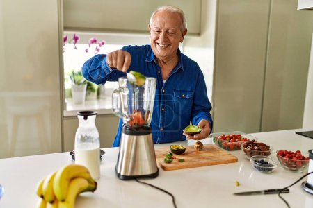 Foto de Hombre mayor sonriendo confiado poniendo aguacate en la licuadora en la cocina - Imagen libre de derechos