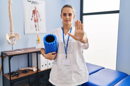 Foto de Young physiotherapist woman holding foam roll at the clinic with open hand doing stop sign with serious and confident expression, defense gesture - Imagen libre de derechos