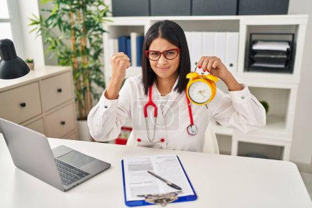 Foto de Young hispanic doctor woman holding alarm clock at the clinic annoyed and frustrated shouting with anger, yelling crazy with anger and hand raised - Imagen libre de derechos