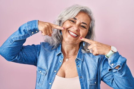Téléchargez les photos : Femme d'âge moyen avec les cheveux gris debout sur fond rose souriant joyeuse montrant et pointant avec les doigts dents et la bouche. concept de santé dentaire. - en image libre de droit