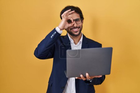 Photo for Handsome latin man working using computer laptop doing ok gesture with hand smiling, eye looking through fingers with happy face. - Royalty Free Image