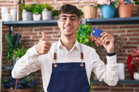 Foto de Joven hombre hispano trabajando en floristería sosteniendo tarjeta de crédito sonriendo feliz y positivo, pulgar hacia arriba haciendo excelente y signo de aprobación - Imagen libre de derechos