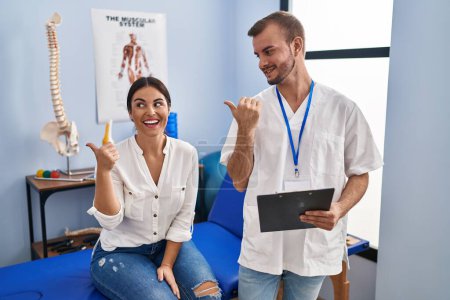 Foto de Young hispanic woman at physiotherapist appointment smiling with happy face looking and pointing to the side with thumb up. - Imagen libre de derechos