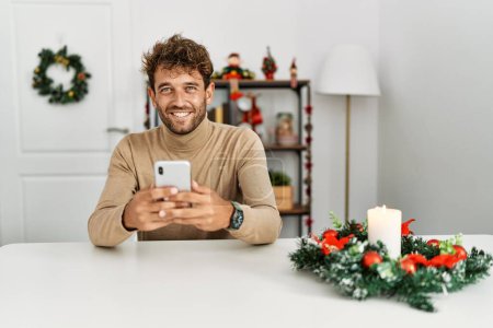 Photo for Young hispanic man using smartphone sitting by christmas decor at home - Royalty Free Image