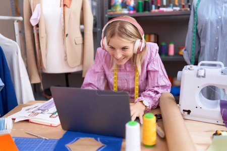 Téléchargez les photos : Jeune femme blonde tailleur utilisant ordinateur portable et écouteurs assis sur la table au magasin de tailleur - en image libre de droit
